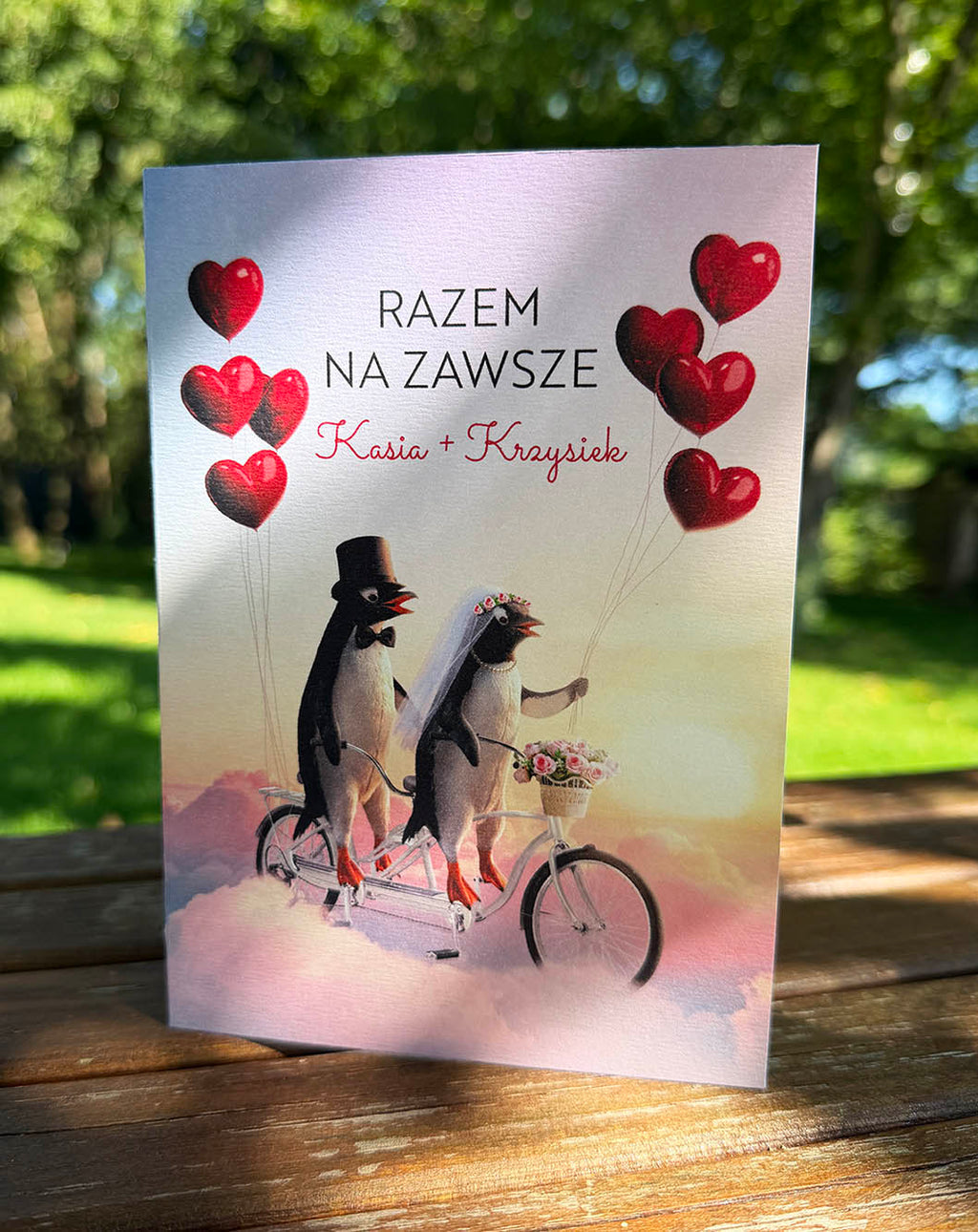 Kartka z życzeniami ślubnymi „Razem na zawsze” przedstawiająca zakochaną parę pingwinów na tandemie, otoczoną chmurami i balonami sercami.